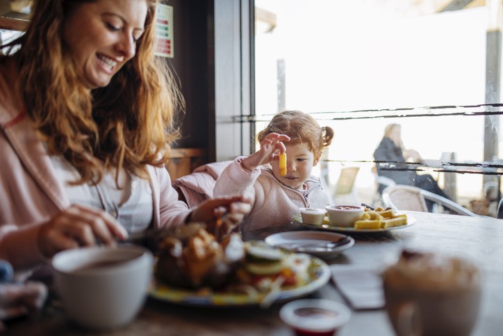 Young family enjoying food at a cafe. The mother is enjoying her food. The little girl is looking at a chip she is holding